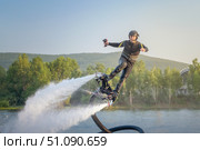 A flyboarding athlete soars above a tranquil lake during the golden hour, showcasing remarkable skills and gravity-defying maneuvers. The backdrop features lush green trees and distant hills, Стоковое фото, фотограф Владимир Разгуляев / Фотобанк Лори