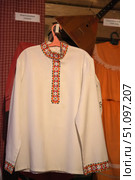 Traditional Russian folk shirt of white color, decorated with embroidery. The embroidery is done in red-black-orange tones and is located along the stand-up collar, on the chest and cuffs. The ornament is a geometric pattern with elements of crosses and diamonds, typical for Slavic motifs. The shirt hangs on a hanger against the background of the interior of a museum or ethnographic exhibition, where a balalaika and other elements of folk life are also visible. The product symbolizes the traditions, culture and originality of the Russian people. Стоковое фото, фотограф Анна Кривицкая / Фотобанк Лори