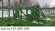 An outdoor sports ground with obstacle course elements. In the center of the frame are two massive metal and wood structures, painted green with a camouflage pattern. Each structure has square holes cut out to imitate windows, and on the side are ladders for climbing. These elements are intended for obstacle course training, physical training, or military sports. In the foreground are visible chains and bars for exercises, and in the background are other elements of the obstacle course. The ground is covered with artificial turf with remnants of snow. The background is a metal fence and bare winter trees. The atmosphere of the picture is emphasized by the combination of outdoor sports, winter season, and the military style of the exercise equipment. Стоковое фото, фотограф Анна Кривицкая / Фотобанк Лори