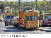 A photo of a historic yellow G-67 service tram in St. Petersburg. The tram is attached to platforms with red sides, decorated with black, orange, white and blue balloons. The photo was taken during a festive parade of retro trams in the city center. In the background, you can see the movement of public transport: buses, a truck and cars. The atmosphere of the photo combines the historical flavor of retro technology and the modern urban rhythm. (2015 год). Редакционное фото, фотограф Анна Кривицкая / Фотобанк Лори