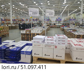 Interior of a large wholesale hypermarket. A spacious sales area with a high ceiling and powerful lighting is filled with rows of pallets with goods. In the center of the frame are packs of A4 office paper of different brands, in particular VOTOCOPY and Mondi IQ Economy. The paper is packed in boxes and stacks on wooden pallets. The price tags indicate the cost - 229.99 rubles for a pack of 500 sheets. In the background, you can see racks with other categories of goods, such as food, household chemicals, hygiene products and office supplies. The atmosphere of the photo reflects a large warehouse-shop with self-service. (2019 год). Редакционное фото, фотограф Анна Кривицкая / Фотобанк Лори