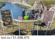 Summer picnic on high bank of the beautiful Belaya river. Tourist table, folding chairs, thermal bag with snacks and thermos on background of riverside on sunny August day. Acive recreation and outdoors. Стоковое фото, фотограф Виктория Катьянова / Фотобанк Лори
