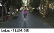 Rear view of young woman jogging along shady paved alley in park wearing grey sportswear with pink accents and sneakers on sunny morning with trees benches. Woman is engaged in sports and fitness. Стоковое видео, видеограф Сергей Тимофеев / Фотобанк Лори