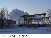 Winter industrial landscape. The frame shows an industrial zone with a tall chimney, from which thick clouds of steam or smoke rise, creating impressive clouds against the background of a brightening sky. In the foreground, snow-covered trees and bushes, a snow-covered road, and metal structures of a manufacturing plant are visible. The atmosphere combines the harsh beauty of winter with industrial power: the whiteness of the snow and the cold shades of the sky contrast with the gray clouds of emissions and the yellow metal of the crane structures. Стоковое фото, фотограф Анна Кривицкая / Фотобанк Лори