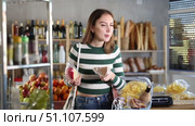Smiling young girl with shopping basket trying to make choice holding two sorts of cheese in grocery store with varied stock. Стоковое видео, видеограф Яков Филимонов / Фотобанк Лори