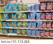 Shelves in a supermarket with a range of products for pets. Large bags of cat litter of different brands and colours are lined up on the shelves. On the left are blue PrettyCat bags with a capacity of 8 litres, in the centre are green Cat Step packages with a picture of a kitten and a green tea aroma, next to them are blue CC Cat bags, and on the right are purple Cat Step with a ginger cat on the package. Prices are posted on white price tags under each shelf. At the top, part of an advertising banner is visible with the inscription "pet in the house". The scene illustrates pet products in retail, a selection of cat litter and the design of the store. (2019 год). Редакционное фото, фотограф Анна Кривицкая / Фотобанк Лори