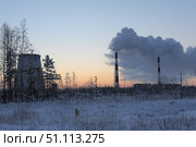 Winter industrial landscape. In the foreground is a snow-covered field with dry grass and thin trees covered with frost. On the horizon rise the chimneys of a large power plant or industrial enterprise, from which thick clouds of smoke rise. On the left is a massive structure similar to an old cooling tower or part of an industrial complex. The sky is painted in soft shades of sunset or sunrise - from pale blue to orange, creating a contrast between the beauty of nature and the heavy impact of industry. The atmosphere of the frame conveys the theme of ecology, energy, industrial production and human impact on the environment. Стоковое фото, фотограф Анна Кривицкая / Фотобанк Лори