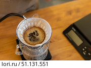 A coffee dripper sits on a scale, with freshly brewed coffee dripping into a glass carafe. The scene features a wooden table and a digital scale. Стоковое фото, фотограф PAv / Фотобанк Лори