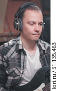 A musician is deeply focused while wearing headphones and holding a black guitar, dressed in a stylish plaid shirt over a white tee, set in a cozy studio environment. Стоковое видео, видеограф Воловодов Владимир / Фотобанк Лори