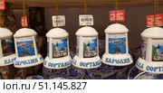 Souvenir ceramic bells with images of Karelia's landmarks, in particular the city of Sortavala. Each bell is decorated with colorful pictures - an Orthodox church with blue domes, landscapes with lakes, rocks and pine trees, as well as images of wildlife, such as a bear. The inscription "Karelia Sortavala" is written at the bottom of the bells. The souvenirs are suspended on golden cords, and price tags are visible at the top. The background is wooden elements and other goods with national motifs. The photo conveys the atmosphere of tourist souvenirs, folk art and regional identity. (2019 год). Редакционное фото, фотограф Анна Кривицкая / Фотобанк Лори