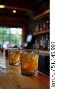 Inviting close-up of condensation-covered glass of amber drink with ice, lined up on rustic wooden bar, capturing warm ambiance of traditional pub with blurred background elements like bottles and nat. Стоковое AI-изображение, AI-художник Данила Большаков / Фотобанк Лори
