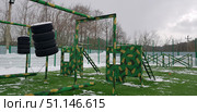 Military sports ground with an outdoor obstacle course in winter. The structures are painted in green camouflage with brown and black spots, imitating the military style. The site has suspended tires, wooden walls with holes, ladders for climbing and crossbars for training. The area is fenced with a high fence, on the ground there is an artificial green lawn, partially covered with snow. In the background you can see bare trees and a cloudy winter sky. This location is used for military training, training teenagers in military-patriotic clubs or for sports games of endurance and agility. Стоковое фото, фотограф Анна Кривицкая / Фотобанк Лори