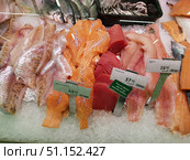 Display cases in the fish section of a supermarket with a variety of fresh fish and fillets laid out on ice. In the foreground are different types of fillets: white fish on the left, bright orange salmon fillet in the center, red tuna fillet, pike fillet on the right. The price tags show the cost: salmon fillet - €24.50/kg, tuna fillet - €37.10/kg, pike fillet - €16.90/kg. In the background, whole fish and a container with small fish (similar to sprats or small herring) are visible. The display case is neatly decorated, the products are placed on a layer of ice to preserve freshness. The image conveys the atmosphere of the store and the choice of seafood for customers. (2019 год). Редакционное фото, фотограф Анна Кривицкая / Фотобанк Лори