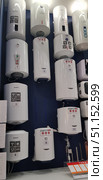 Display cases of a home appliance store with various models of electric water heaters. On the wall, storage water heaters of different volumes and capacities are placed in several rows. The bodies are mostly white, cylindrical and rectangular in shape. Some models are equipped with informative stickers with characteristics: volume in liters, power in kilowatts, type of tank coating. The brands Ariston, EDISSON and others are visible. In the foreground are price tags and an information stand about heating elements. The scene conveys the atmosphere of a home appliance store and demonstrates the assortment for customers to choose from. (2018 год). Редакционное фото, фотограф Анна Кривицкая / Фотобанк Лори