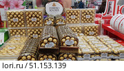 In a large supermarket or hypermarket. In the foreground is a bright and spectacular display of Ferrero Rocher chocolates in branded gold foil. The chocolates are packed in transparent plastic boxes of different formats - rectangular and oval. The Ferrero Rocher brand logo is visible on the packaging. The gold packaging creates an atmosphere of celebration, luxury and premium. In the background is a sales area with other customers, shelves and advertising structures. The photo conveys the atmosphere of pre-holiday shopping, abundance and choice of sweets. (2019 год). Редакционное фото, фотограф Анна Кривицкая / Фотобанк Лори