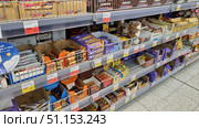 A shelf in a supermarket with a large selection of chocolate bars and sweets. The assortment includes popular brands - Twix, Snickers, Milka, Picnic, Mars and others. The bars are laid out in open boxes and metal baskets, price tags are attached to the shelves. The photo shows several levels with a variety of sweet goods: chocolate, wafers, candies and nut mixes. The atmosphere of a typical grocery store, illustrating the theme of shopping, sweets and everyday food choices. (2021 год). Редакционное фото, фотограф Анна Кривицкая / Фотобанк Лори