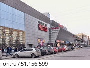 Modern shopping mall "Voyage" in the urban development. The building is made in a modern architectural style with cladding made of gray panels and large mirrored windows, which reflect residential buildings. On the facade there are signs of the cinema "Mireille", restaurants, shops and chain brands, among which McDonald's is visible. In front of the shopping center there is a parking lot with parked cars and pedestrians walking along the building. The scene conveys the atmosphere of everyday city life, the convenience of shopping malls and the dynamics of the metropolis. (2015 год). Редакционное фото, фотограф Анна Кривицкая / Фотобанк Лори