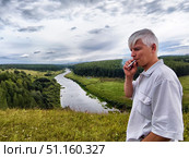 Man enjoying a cigarette on a cliff overlooking a river surrounded by lush green forest on a cloudy day. Стоковое фото, фотограф Кривошеина Елена Леонидовна / Фотобанк Лори