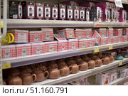 Shelves in a shop with kitchenware. In the foreground are brown ceramic teapots with lids and handles, neatly arranged in rows. Above are boxes with teapots in red and white checkered packaging with an image of a teapot. On the top shelf you can see French presses for coffee and tea in black and pink boxes. The shelves are well lit, the goods are displayed in an orderly manner, creating an atmosphere of a large selection of utensils for making hot drinks. This image would be suitable for illustrating household goods stores, kitchenware stores and supermarkets. (2015 год). Редакционное фото, фотограф Анна Кривицкая / Фотобанк Лори