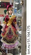 Decorative doll in a national costume. The doll is dressed in a bright burgundy dress with rich embroidery, gold decor and green inserts of imitation stones. The skirt is decorated with patterns with elements of folk ornaments, lace and frills made of white and lilac fabric. On the doll's head is a burgundy headdress, complemented by a gold headband and decorations. The costume is detailed: there are wide sleeves made of transparent fabric with a gold pattern, as well as decorative accessories that emphasize the traditional style. The doll is behind the glass of the display case, other products and souvenirs are visible nearby.The image is perfect for illustrations related to ethnography, folk art, culture, national clothing, traditional crafts and souvenirs. Стоковое фото, фотограф Анна Кривицкая / Фотобанк Лори
