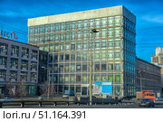 A modern office building in the style of Soviet modernism, located in St. Petersburg. The architecture of the building is characterized by strict geometric forms, the facade is made entirely of rectangular glass sections in metal frames. In the foreground, street lamps, wires and billboards are visible. Cars are driving along the road, including a truck and cars. The building stands out for its large size and the mesh structure of the facade, reflecting the blue sky. Below are shop windows and signs. The atmosphere of the frame conveys the everyday city life of St. Petersburg. (2019 год). Редакционное фото, фотограф Анна Кривицкая / Фотобанк Лори