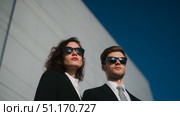 A stylish man and woman in black suits and sunglasses stand confidently against a modern building and a vivid blue sky. Their serious expressions and sharp formal attire give them a secret agent or security professional look. This image can be interpreted in connection with Defender of the Fatherland Day (February 23) or National Unity Day (November 4) in Russia, symbolizing strength, duty, and patriotism. Стоковое AI-изображение, AI-художник Ксения Чунаева / Фотобанк Лори