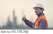 Smiling industrial worker in a protective uniform and hard hat showing thumbs up outdoors with refinery plant in the background. Concept of industry, safety, oil and gas production, engineering professions. Suitable for the celebration of Oil and Gas Industry Workers Day in Russia. Стоковое AI-изображение, AI-художник Ксения Чунаева / Фотобанк Лори