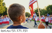 A child stands on the sidewalk with flags, enjoying a colorful parade filled with excitement and patriotism. Стоковое AI-видео, аниматор Данила Большаков / Фотобанк Лори