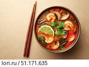 Bowl of Tom Yum Shrimp and Mushroom hot soup, garnished with fresh coriander leaves, herbs, lime slices, chili peppers. Beige table background, chopsticks pair. Traditional Thai Prawn Tom Yum Soup. Стоковое AI-изображение, AI-художник Марина Демидюк / Фотобанк Лори