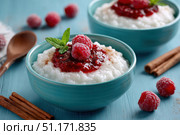 Milky Rice pudding with superberries and red raspberry sauce, served in a white bowl on blue tablecloth background. Rice porridge topped with some raspberries and cinnamon sprinkled. Old English style. Стоковое AI-изображение, AI-художник Марина Демидюк / Фотобанк Лори