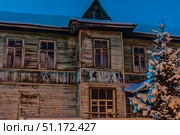 An old wooden house on a winter evening. The two-story building with peeling paint on the facade and old wooden frames looks abandoned or forgotten by time. The walls are covered with traces of moisture and darkening, which emphasizes the age of the building. In the foreground, a spruce tree with branches covered with fluffy snow is visible, which creates a contrast between the cold winter beauty and the gloomy atmosphere of the old house. The deep blue sky enhances the feeling of night and silence. The photo conveys an atmosphere of mystery, history and the elusive past. Стоковое фото, фотограф Анна Кривицкая / Фотобанк Лори
