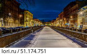 Winter St. Petersburg in the evening. The picture shows a snow-covered frozen canal, on both sides of which are lined with old houses in the classical and modern styles. The cozy cityscape is illuminated by yellowish lanterns, creating a warm contrast with the cold blue of the winter sky. Cars are parked along the embankment, and city lights are visible in the distance. The atmosphere of the picture conveys the tranquility, romance and grandeur of a northern city famous for its architecture and embankments. Стоковое фото, фотограф Анна Кривицкая / Фотобанк Лори