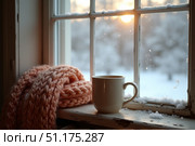 A warm cup rests beside a soft, pink scarf on a windowsill, framed by frosty windowpanes. Outside, snow blankets the ground, illuminated by the gentle light of dawn. Стоковое AI-изображение, AI-художник Ольга Игнашина / Фотобанк Лори