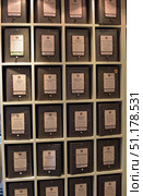 A shelf of a specialized tea shop with metal containers for storing and selling loose tea. Each container has a plate with the name of the tea, description and price per 100 grams. On the plates you can read the names of different varieties: pu-erh, darjeeling, assam, green, milky oolong, Chinese and Indian teas, as well as signature blends. The containers are made in the same style - dark metal boxes with neat labels, located in the cells of a wooden rack. At the bottom of each container there is a dispenser for convenient dispensing of loose tea. The atmosphere of the photo conveys the coziness of a tea shop and the variety of assortment designed for connoisseurs and gourmets. (2015 год). Редакционное фото, фотограф Анна Кривицкая / Фотобанк Лори