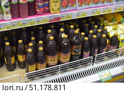 A shelf in a supermarket with drinks. In the center of the frame are numerous dark-colored plastic bottles of kvass with golden caps. Popular brands such as "Russkiy Dar" and "Tsarskie Pripasy" are noticeable among them. The bottles have a characteristic relief pattern in the form of honeycombs. On the top shelf are boxes with juices and drinks in tetra packs, including berry fruit drinks and jelly. On the right, you can see yellow packages with juice. The price tags indicate prices in rubles, and on the bottom shelf, a green sign highlights the inscription "New!" The store lighting emphasizes the gloss of the bottles, creating a feeling of richness and abundance of goods. (2015 год). Редакционное фото, фотограф Анна Кривицкая / Фотобанк Лори
