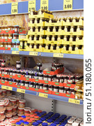 Supermarket shelves filled with various sweet products in jars and packages. At the top of the rack are rows of plastic bottles of honey in yellow boxes with price tags. Just below are jars of jam and preserves of various flavors in glass containers: cherry, blueberry, apricot, strawberry. The jars are organized in cardboard boxes with the Maribel brand logo, creating neat colored rows. At the bottom of the photo you can see jars of peanut butter, creams and other sweet spreads. Yellow price tags with prices in euros and large inscriptions in Finnish indicate that the store belongs to a European retail chain. The atmosphere of the photo conveys the abundance of choice and the organization of the grocery department. (2015 год). Редакционное фото, фотограф Анна Кривицкая / Фотобанк Лори