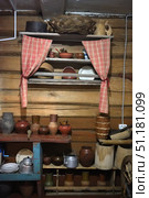 Interior of a traditional Russian village hut with elements of old-fashioned life. On the wooden wall hangs a shelf, covered with red and white checkered curtains, where clay bowls, plates and wooden utensils are placed. At the top of the shelf lies a wicker basket and a wooden jug. Below are various objects: clay pots and jugs of different shapes and sizes, an aluminum teapot, wooden troughs, spoons and wicker baskets. The interior feels like a genuine atmosphere of rural life, filled with details of a traditional peasant kitchen. Wooden walls and shelves emphasize the ethnographic value of the objects, creating a sense of authenticity and returning to the origins of Russian life. Стоковое фото, фотограф Анна Кривицкая / Фотобанк Лори