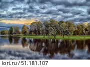 A picturesque landscape with trees on the river bank in the evening. The water reflects the treetops and the dramatic sky covered with thick gray clouds. On the horizon, you can see the soft orange-yellow glow of the sunset, creating a contrast with the cool shades of the sky and water. The bank is covered with green grass, and the trees standing by the water form a harmonious line, emphasizing the calm and beauty of nature. The atmosphere of the photo conveys a combination of peace and light drama, characteristic of the pre-sunset time. Стоковое фото, фотограф Анна Кривицкая / Фотобанк Лори