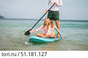 Father enjoys stand up paddleboard with kids on summer vacation. Happy children sit on board, smiling and playing while parent paddles in calm sea. Concept of outdoor activity and family fun. Стоковое фото, фотограф Енваев Андрей / Фотобанк Лори