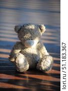 A grey teddy bear from the Me to You collection. The toy has a recognizable blue nose, patches on its paws and head, which gives it a touching and slightly vintage look. The bear sits on a wooden surface illuminated by soft warm sunlight, through which long shadows fall. The atmosphere of the frame creates a feeling of coziness, tenderness and slight nostalgia. Such a photo is perfect for the theme of childhood, gifts, romance, love, sentimentality and the symbolism of friendship. Стоковое фото, фотограф Анна Кривицкая / Фотобанк Лори