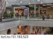 The photo was taken inside a modern shopping center. In the foreground, two levels of the building are visible with spacious corridors decorated with green plants. On the upper floor, there is an interactive navigation screen with a map of the shopping center, and next to it stands a woman in a bright yellow jacket. In the center of the frame is the entrance to the LINDEX store with display cases and bright lighting, and part of the same store is also visible on the lower level. Shoppers walk along the corridors, creating an atmosphere of a lively shopping space. The interior is designed in a modern style with elements of glass partitions and metal structures, which emphasizes the style and spaciousness of the building. (2015 год). Редакционное фото, фотограф Анна Кривицкая / Фотобанк Лори