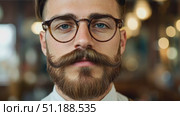 Portrait of a young man with a styled beard and mustache, wearing round eyeglasses and a white shirt. His well-groomed appearance and confident gaze convey a hipster aesthetic, blending classic grooming with modern fashion. The blurred background with warm lighting suggests a cozy café or barbershop setting, emphasizing themes of masculinity, personal style, and urban culture. Стоковое AI-видео, аниматор Ксения Чунаева / Фотобанк Лори