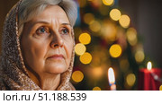 A pensive elderly woman in a headscarf gazes into the soft glow of a candle, framed against warm festive lights in the background. The scene evokes a deep spiritual and emotional atmosphere, commonly associated with Orthodox Christian holidays such as Christmas or Easter, or days of solemn national remembrance in Russia. This quiet moment of reflection captures the essence of faith, tradition, and memory, particularly resonant during Orthodox memorial services or cultural observances like the Day of Remembrance and Sorrow. It symbolizes the intersection of personal devotion, generational memory, and Russian spiritual heritage. Стоковое AI-видео, аниматор Ксения Чунаева / Фотобанк Лори