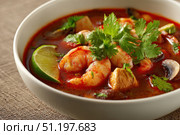 Bowl of Tom Yum Shrimp and Mushroom hot soup, garnished with fresh coriander leaves, herbs, lime slices, chili peppers. Beige table background, chopsticks pair. Traditional Thai Prawn Tom Yum Soup. Стоковое AI-изображение, AI-художник Марина Демидюк / Фотобанк Лори
