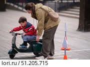 Woman assisting a young boy riding a toy scooter on a wooden boardwalk. Mother helping child learn riding skills, teaching concept. A child aged 3 years. Стоковое фото, фотограф Андрей Журавлев / Фотобанк Лори