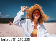 Happy woman in straw hat and sunglasses laughing while relaxing on sandy beach. Concept of joy, carefree spirit, and glowing holiday lifestyle. Стоковое фото, фотограф Енваев Андрей / Фотобанк Лори