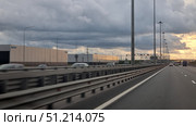 Section of the Saint Petersburg Ring Road (KAD). The wide multi-lane highway stretches into the distance, with lampposts on both sides. There are cars moving on the road, trucks and cars are visible. Large industrial buildings and warehouses with company logos are located on the left. Road structures and overpasses are visible in the distance. The sky is covered with thick gray clouds, but the sunset light breaks through on the horizon, creating a beautiful contrast between the dark clouds and the golden reflections on the facades of the buildings and in the sky. The atmosphere of the photo combines the dynamics of movement and the urban industrial landscape. Редакционное фото, фотограф Анна Кривицкая / Фотобанк Лори