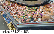 A display refrigerator in a supermarket filled with various types of sausages. The display case includes cooked sausages, frankfurters, wieners, smoked and semi-smoked sausages, as well as sliced ​​sausages in vacuum packaging. The products are neatly laid out: in the foreground, sausages and wieners in film are visible, behind them are loaves of cooked sausage and packages with slices. The packages have bright green, yellow and red labels. Under the glass are price tags with current prices for the products. At the bottom of the photo, glass jars of mustard and packages of bottled drinks are visible. The atmosphere of a typical supermarket with a large selection of meat products. Редакционное фото, фотограф Анна Кривицкая / Фотобанк Лори