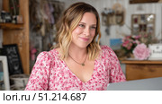 A cheerful woman in a floral pink blouse sits comfortably in what appears to be a cozy, creatively decorated boutique or home workspace. Her warm smile and relaxed posture exude friendliness and approachability. The soft lighting and pastel-toned background, adorned with flowers and personal decor, suggest a welcoming environment, possibly a small business, studio, or artistic space. This setting and expression evoke themes of entrepreneurship, creativity, customer service, and modern femininity. The image aligns well with ideas of small business ownership, lifestyle branding, and female-led creative enterprises. Стоковое AI-изображение, AI-художник Ксения Чунаева / Фотобанк Лори