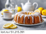 Homemade lemon drizzle cake with icing. Classic lemon pound cake with citrus glaze and zest. Whole round, side view, closeup. Fresh homemade baked dessert with lemon slices. Blurred tea set background. Стоковое AI-изображение, AI-художник Марина Демидюк / Фотобанк Лори